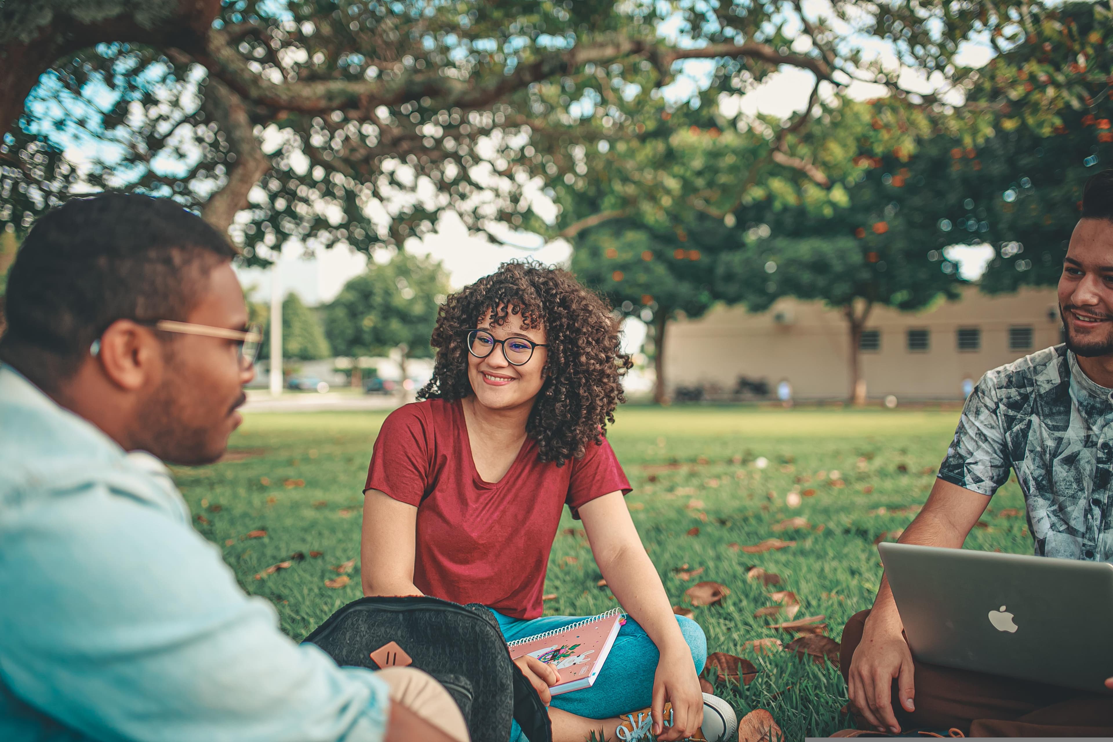 Three university students smiling and studying together outdoors on a sunlit campus lawn